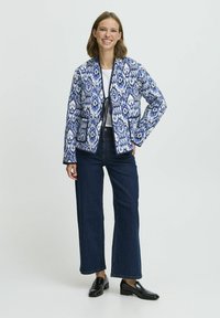 Quiltrad jacka i blå och vit ikatmönster, knäppning framtill, två fickor fram, bärs över en vit topp och mörkblå jeans, med svarta skor.