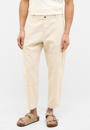 Beige Cargo-Hosen mit lockerer Passform, ausgestattet mit vier Taschen und sichtbaren Nähten. Hergestellt aus strapazierfähigem Baumwollstoff. Kombiniert mit hellbraunen Sandalen.