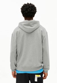 Grijze capuchontrui met een losse pasvorm, geribbelde boorden en zoom. Draagt over een blauwe gelaagde blouse en heeft een label op de zwarte broek.