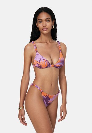 Donna con capelli lunghi e neri indossa un bikini floreale viola e arancione con spalline arricciate, orecchini a cerchio dorati e un braccialetto d'oro.