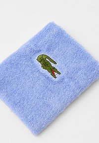 Lyseblå frottéhåndkle med en teksturert overflate og en brodert grønn krokodillelogo på den ene siden.