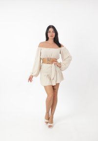 Beige Off-Shoulder-Crop-Top mit langen, voluminösen Ärmeln, kombiniert mit einem passenden Minirock mit seitlichem Bindeband. Glattes, strukturiertes Material.