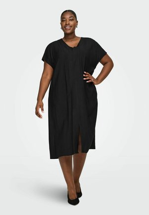 Smilende kvinde stående med hånd på hoften iført en løs sort midi-kjole og sorte hæle mod en ensfarvet baggrund.