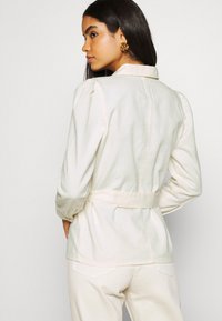 Giacca leggera color crema con cintura, maniche puffed, design strutturato e dettaglio sulla spalla posteriore. Realizzata in tessuto traspirante, presenta una sottile texture.