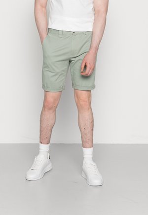Hombre vistiendo pantalones cortos de color verde claro con dobladillo enrollado, calcetines blancos y zapatillas blancas, de pie contra un fondo liso y claro.