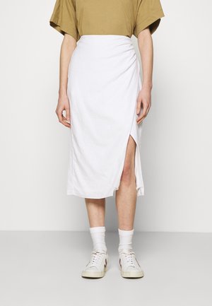 Person iført en hvid midi-nederdel med en slidse foran, beige kortærmet top, hvide sokker og hvide sneakers.