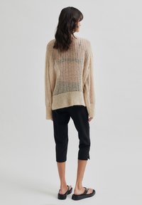 Pull en maille beige léger à côtes, présentant un tissage lâche, des manches longues et une coupe décontractée. Associé à un pantalon noir court et des sandales plates.