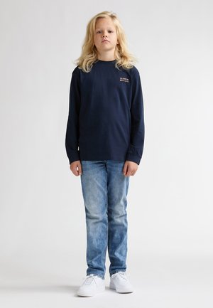 Dunkelblaues Langarm-T-Shirt mit kleinem aufgedrucktem Logo, kombiniert mit hellblauen Jeans und weißen Sneakern vor einem schlichten Hintergrund.