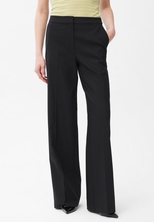 Kobieta ubrana w czarne spodnie z szerokimi nogawkami i wysokim stanem z bocznymi kieszeniami, w połączeniu z jasnożółtym, bezrękawnym topem oraz czarnymi, spiczastymi butami.