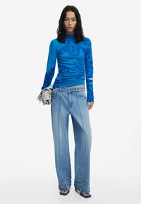 Blaues Langarmoberteil mit floralem Muster, kombiniert mit hellblauen weiter geschnittenen Jeans. Als Accessoire dient eine gedruckte Stofftasche, die in der Hand gehalten wird.