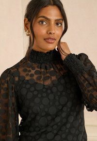 Crna prozirna majica s velikim kružnim uzorcima, visokim nabranim ovratnikom i elastičnim manžetama. Mekana tekstura, lagana tkanina i uski kroj.