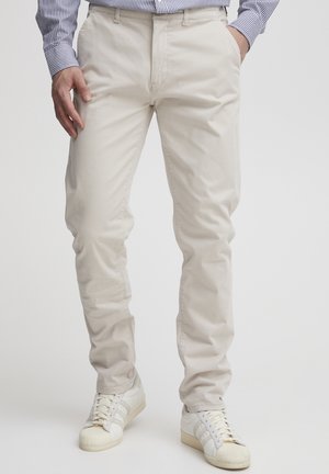 Mężczyzna w beżowych spodniach, białych trampkach i niebieskiej koszuli w paski, stojący z rękami w kieszeniach na jednolitym tle.