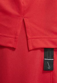 Red athletic shirt with textured fabric, featuring a split hem and a black tag with a white Nike logo. Flat seams and short sleeves.