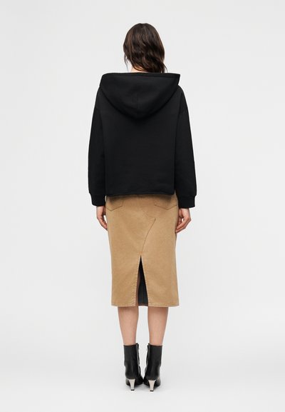 Γυναίκα που στέκεται γυρισμένη προς τα πίσω φορώντας μαύρο φούτερ με κουκούλα, μπεζ midi φούστα με σκίσιμο στο πίσω μέρος και μαύρα μποτάκια με τακούνι, μπροστά σε λευκό φόντο.