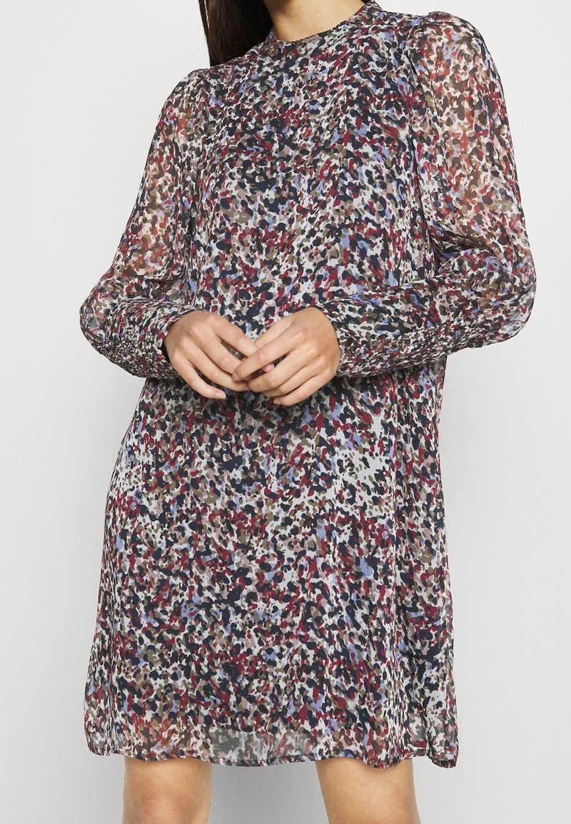 Femme portant une robe à manches longues imprimée d’un motif abstrait multicolore avec des poignets froncés, debout devant un fond uni.