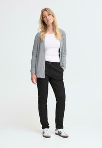 Woman standing with one hand in pocket, wearing gray cardigan, white shirt, black pants, and white sneakers with black stripes.