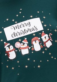 O tecido verde apresenta quatro pinguins de desenho animado a usar chapéus de inverno vermelhos e brancos, segurando um cartaz de "Feliz Natal", com detalhes em estrelas douradas.