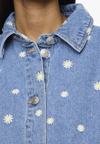 Vaaleansininen farkkutakki, jossa on valkoista ja keltaista päivänkakkara-embroidausta, hopeiset nepparit ja klassinen kaulussuunnittelu.