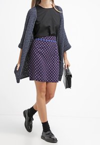 Svart topp, marinblå stickad cardigan, lila kjol med diamantsmönster, svarta strumpor och svarta skor. Håller en svart handväska.