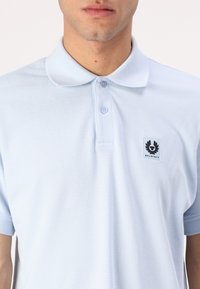 Ανοιχτό μπλε πόλο μπλούζα φτιαγμένη από υφασμάτινο υλικό. Διαθέτει κλασικό γιακά, δύο κουμπιά και μαύρο λογότυπο στην αριστερή πλευρά του στήθους.