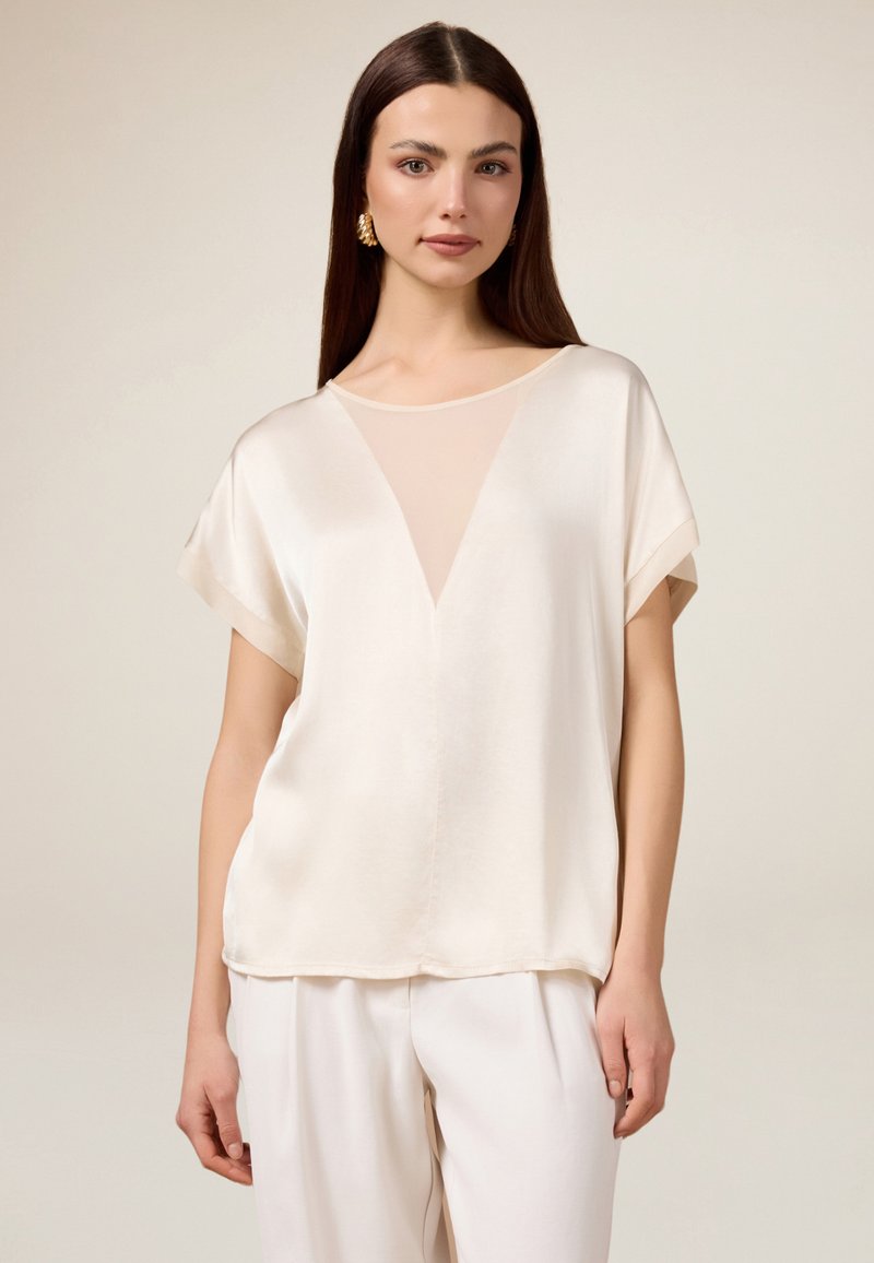 Crème zijden blouse met een doorschijnende inzet en korte mouwen, voorzien van een V-hals ontwerp en een gladde textuur. Gecombineerd met lichtgekleurde broeken.