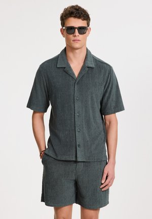 Camisa de manga corta con botones y pantalones cortos a juego en un tejido texturizado de color verde oscuro, con un patrón sutil y un corte relajado.