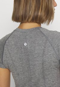 Cheveux bruns mi-longs et partie supérieure du dos d’une personne portant un t-shirt de sport gris ajusté avec un petit logo blanc Lululemon.