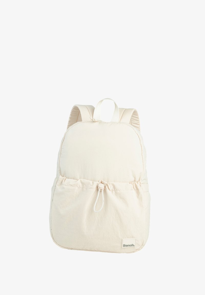 Beige fabric backpack with a rounded shape, padded back, adjustable straps, and a drawstring pocket at the front. Features a logo tag.