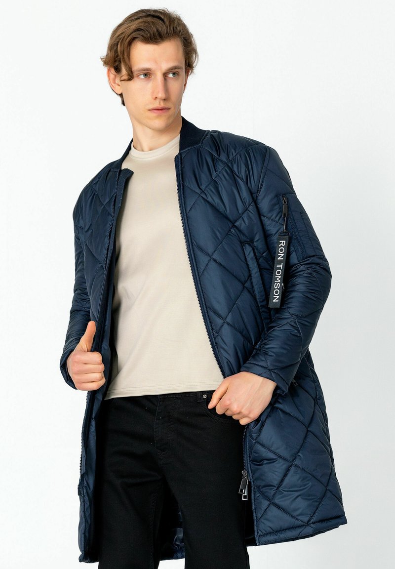 Steppjacke in Marineblau mit Reißverschluss, versehen mit einem schwarzen Etikett am Ärmel, aus einem glänzenden Material gefertigt. Enthält seitliche Taschen.