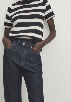 Persoon draagt een zwart-wit horizontaal gestreept T-shirt met korte mouwen en donkerblauwe high-waisted jeans, handen in de zakken.