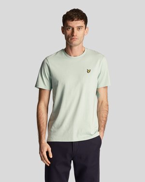 Homme portant un T-shirt à manches courtes vert clair avec un petit logo d'oiseau jaune, un pantalon sombre, se tenant avec une main dans la poche contre un fond gris.