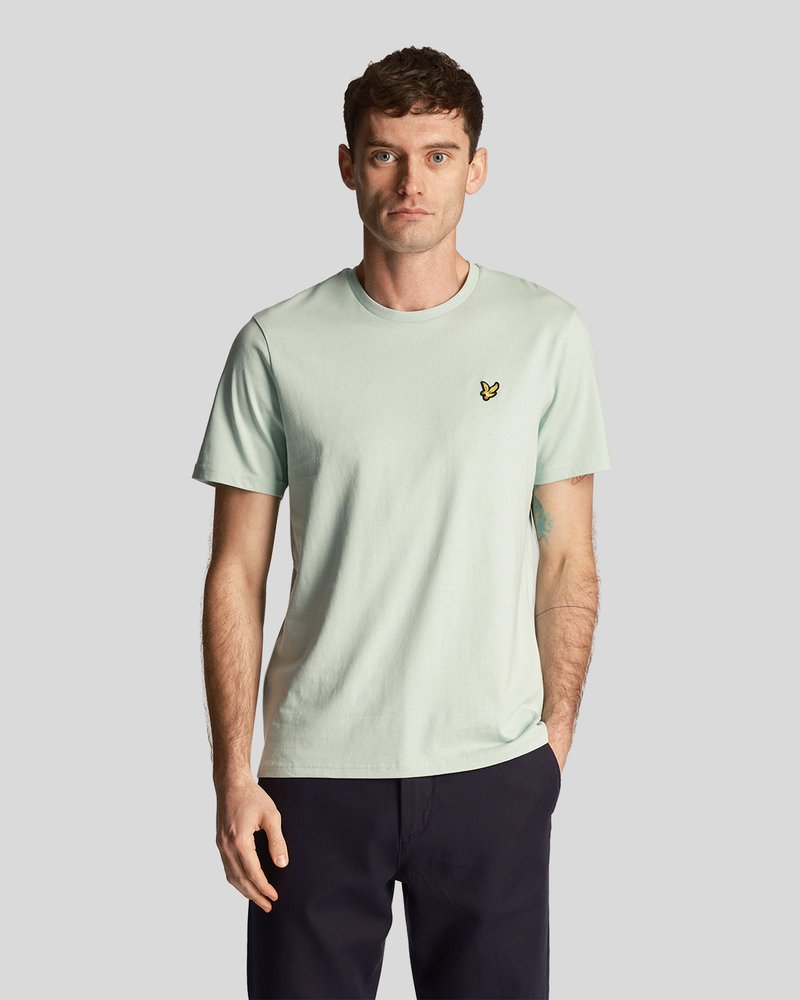 Homme portant un T-shirt à manches courtes vert clair avec un petit logo d'oiseau jaune, un pantalon sombre, se tenant avec une main dans la poche contre un fond gris.