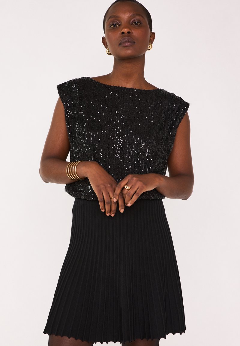 Top senza maniche nero ricoperto di paillettes abbinato a una gonna nera plissettata. L'outfit presenta un tessuto texture e una silhouette aderente.