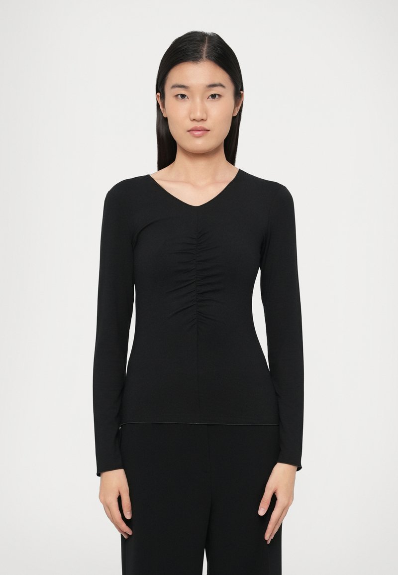 Μαύρη μπλούζα με μακριά μανίκια και λαιμόκοψη V, με λεπτομέρειες πιέτας μπροστά, κατασκευασμένη από απαλό, ελαστικό ύφασμα. Εφαρμοστή γραμμή.