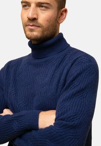 Suéter de cuello alto azul marino hecho de un tejido de punto texturizado, con un patrón ondulado y puños acanalados. Diseño ajustado con un cuello alto.