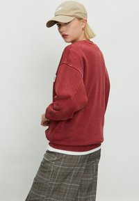 Młoda osoba w beżowej czapce Nike, czerwonym oversize'owym swetrze, białej koszulce i szarych spodni plaid, spoglądająca przez ramię.