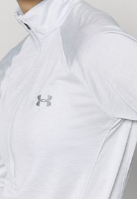 Vaaleanharmaa urheilupulloveri korkealla kauluksella, jossa puolihalkio vetoketju ja teksturoidut olkapaneelit. Pieni logo rinnassa.