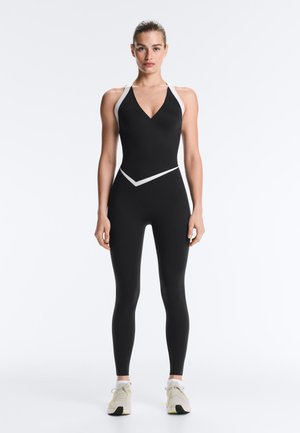 Vrouw staand met het gezicht naar voren, draagt een zwarte mouwloze atletische jumpsuit met witte accenten en witte sneakers tegen een witte achtergrond.