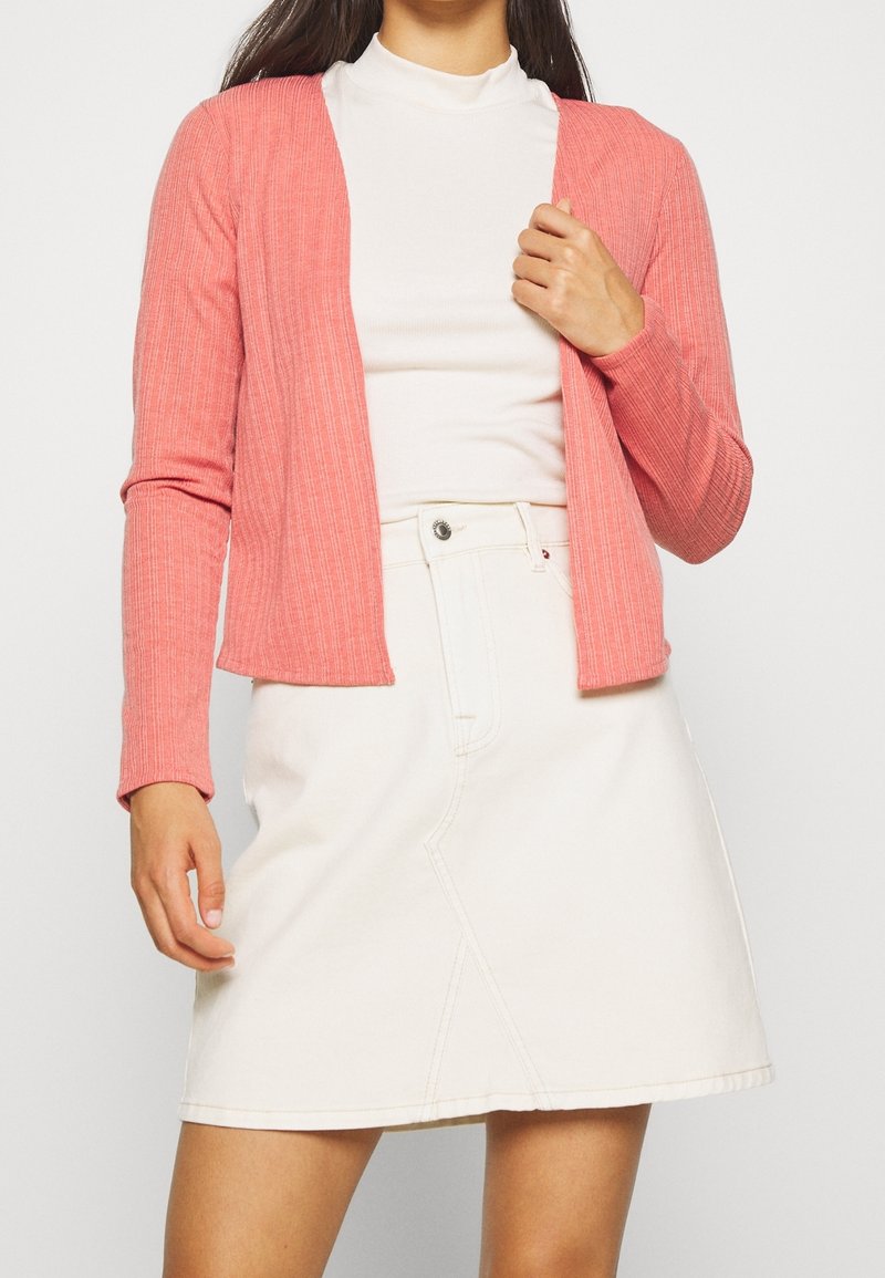 Korálový pletený kardigan s dlouhými rukávy a otevřeným předním designem, kombinovaný s bílou džínsovou sukní. Jednoduchá a texturovaná tkanina s minimem švů.