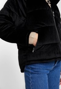 Czarna kurtka puchowa o gładkiej, błyszczącej fakturze, z kieszeniami na zamki i stójką, zestawiona z beżowym swetrem i niebieskimi dżinsami.