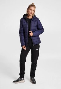 Mörkblå vadderad jacka med luva, dragkedja framtill och detaljerad sömnad. Bärs över en svart topp och matchas med svarta joggingbyxor och sneakers.