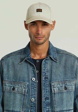 Man draagt een beige pet en een lichtblauwe spijkerjas over een zwart shirt, kijkt neutraal vooruit tegen een effen achtergrond.