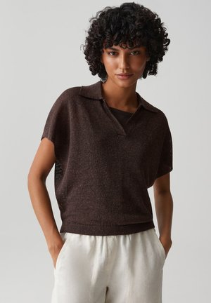 Frau mit kurzen lockigen Haaren, die ein dunkelbraunes kurzärmeliges Strickoberteil und hellbeige Hosen trägt, mit den Händen in den Taschen vor einem schlichten Hintergrund stehend.