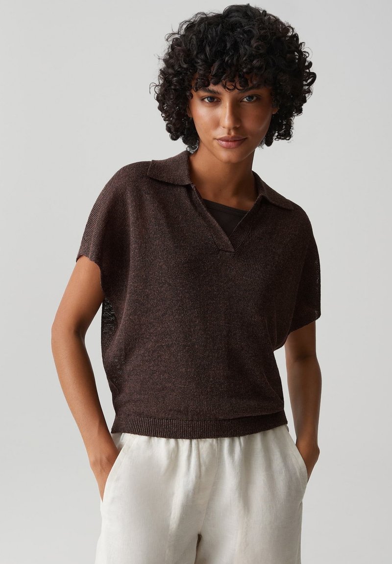 Woman with short curly hair wearing a dark brown short-sleeve knit top and light beige pants, standing with hands in pockets against plain background.