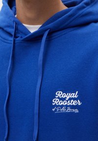 Royalblå hoodie med dragsko på huvan. Har vit broderad text "Royal Rooster" och "el Pollo Bravo" på framsidan. Mjuk textur.