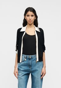 Μαύρο μπουφάν με φερμουάρ και λευκό γιακά με κείμενο λογότυπου, φορεμένο πάνω από μαύρο τοπ, συνδυασμένο με ανοιχτό μπλε τζιν.