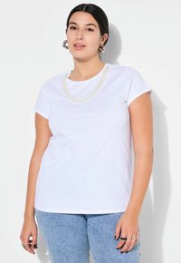 T-shirt bianca in cotone con maniche corte, caratterizzata da una collana di perle a strati e un collo rotondo. Abbinata a jeans azzurri chiari.
