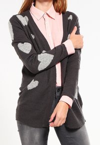 Šedý cardigan s jemnými světle šedými vzory srdíček, otevřeným předním dílem a texturovaným pletením. V kombinaci s bledě růžovou košilí na knoflíky pod ním.