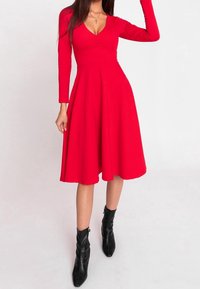 Robe longue à manches longues rouge avec un décolleté en V profond, un corsage ajusté et une jupe évasée. Portée avec des bottines noires, mettant en valeur une texture lisse.