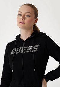 Juodas užtrauktukas gobtuvu, kuriame užrašas "GUESS" blizgančiomis rhinestone detalėmis. Su raišteliais ir efektyviais elastiniais rankogaliais, užtikrinančiais patogų prigludimą.